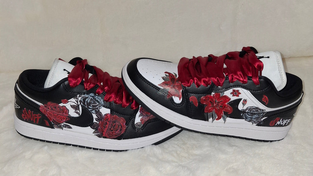 A pair of low-top sneakers with a black and white base, decorated with red roses, red satin-like laces, and red floral graphics. 