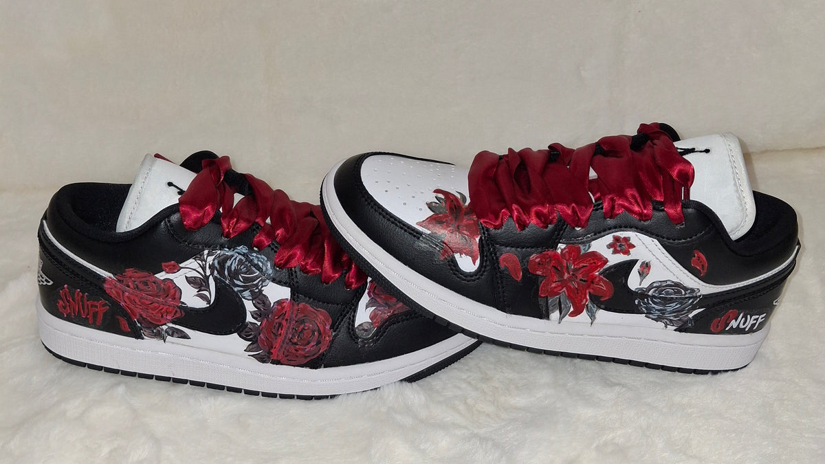 A pair of low-top sneakers with a black and white base, decorated with red roses, red satin-like laces, and red floral graphics. 