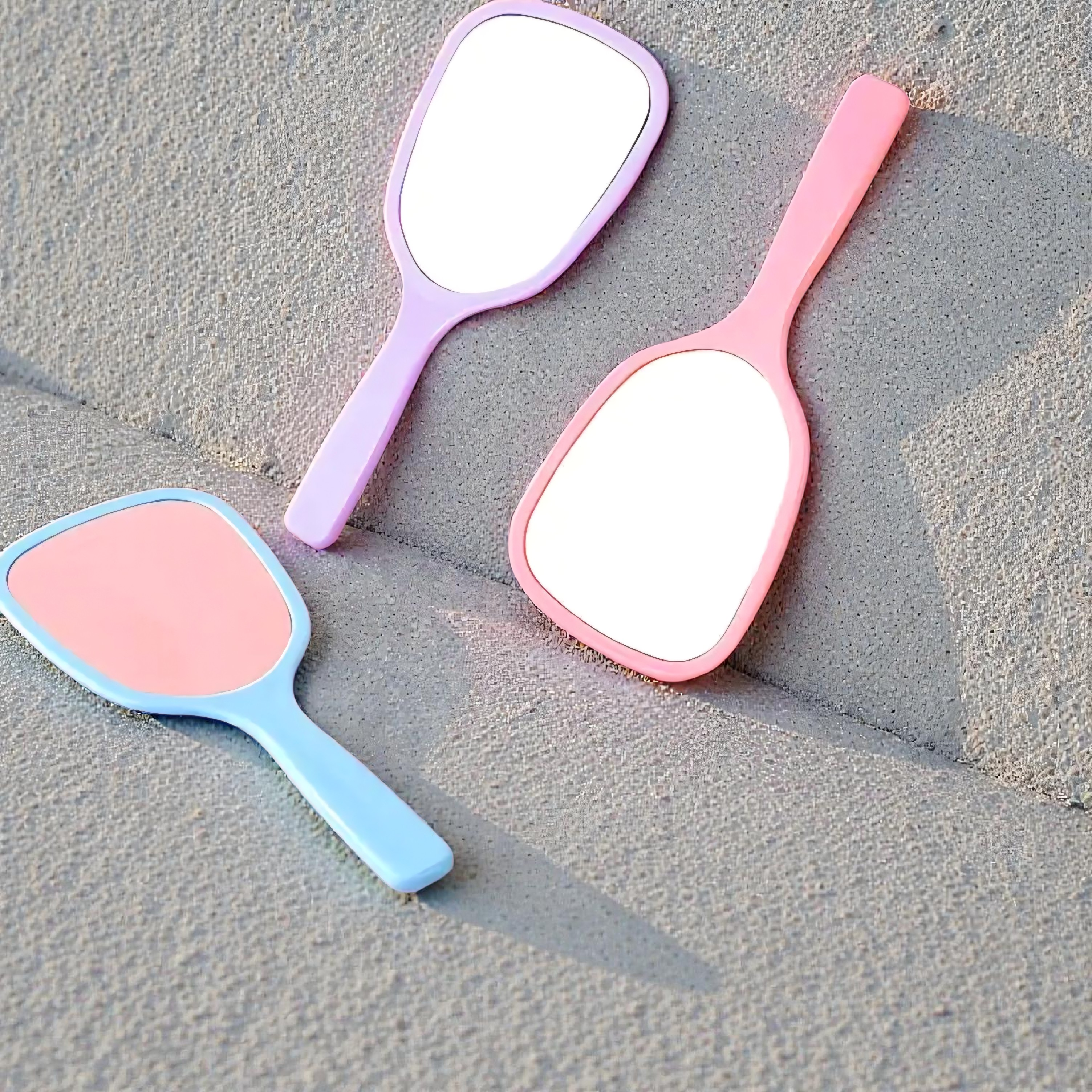 Three handheld mirrors in pink, blue, and purple on a textured surface.